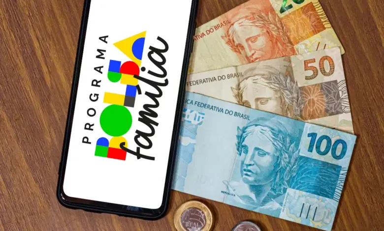 Smartphone exibindo logo Bolsa Família ao lado de notas e moedas de real sobre mesa de madeira.
