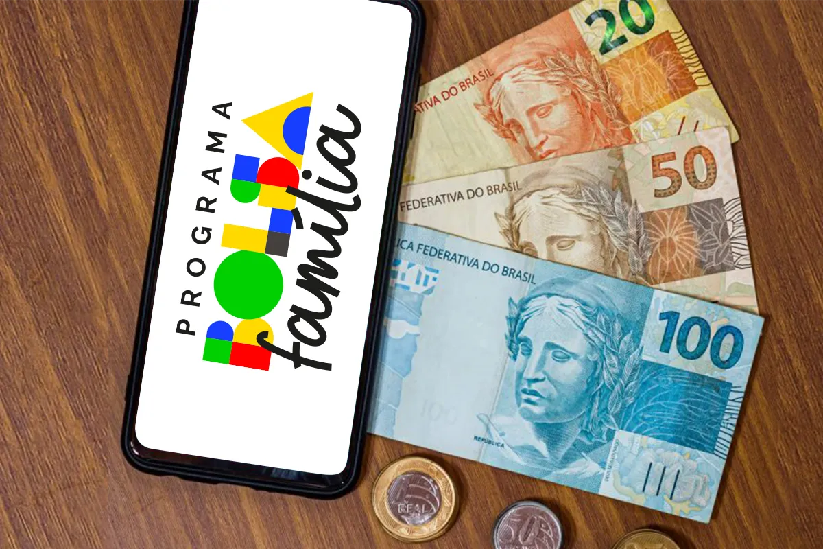 Smartphone exibindo logo Bolsa Família ao lado de notas e moedas de real sobre mesa de madeira.