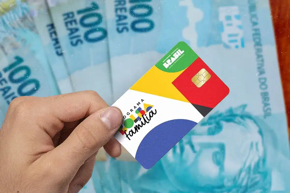 Mão segura cartão do Bolsa Família sobre várias notas de cem reais