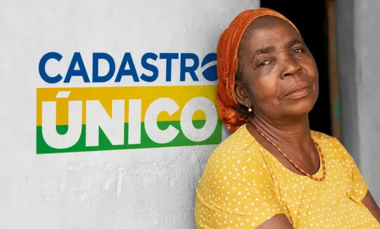 Mulher de lenço laranja e blusa amarela encostada em parede com logo do Cadastro Único ao fundo