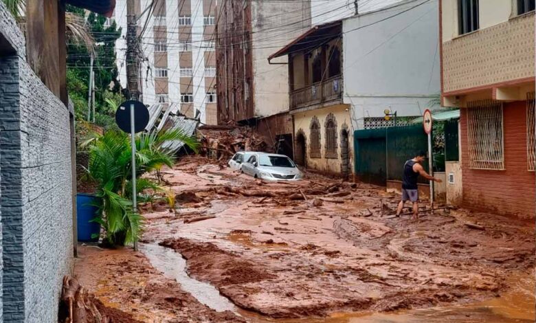 Rua coberta por lama após enchente com carro parcialmente submerso e homem caminhando em meio à destruição
