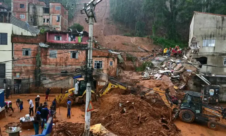 Retroescavadeiras amarelas removem lama de deslizamento em comunidade com casas de tijolos vermelhos enquanto moradores observam.