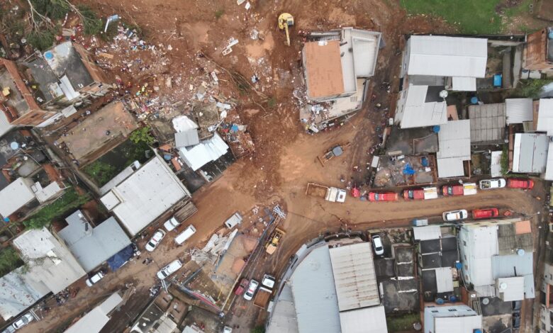 Vista aérea de deslizamento de terra atingindo casas em bairro residencial com escavadeiras e equipes de resgate no local.