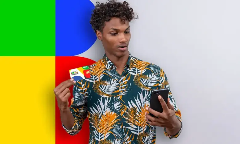 Jovem negro de cabelos cacheados, camisa tropical verde e laranja, segura cartão Bolsa Família e olha celular preto.