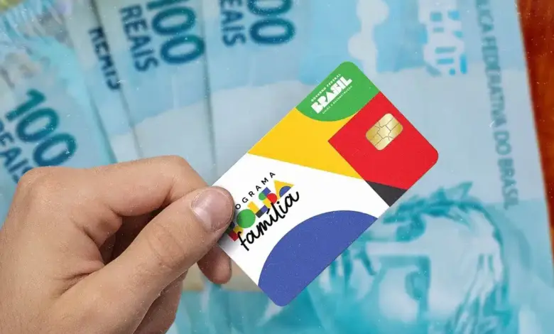 Mão segurando cartão colorido do Bolsa Família com notas de 100 reais ao fundo