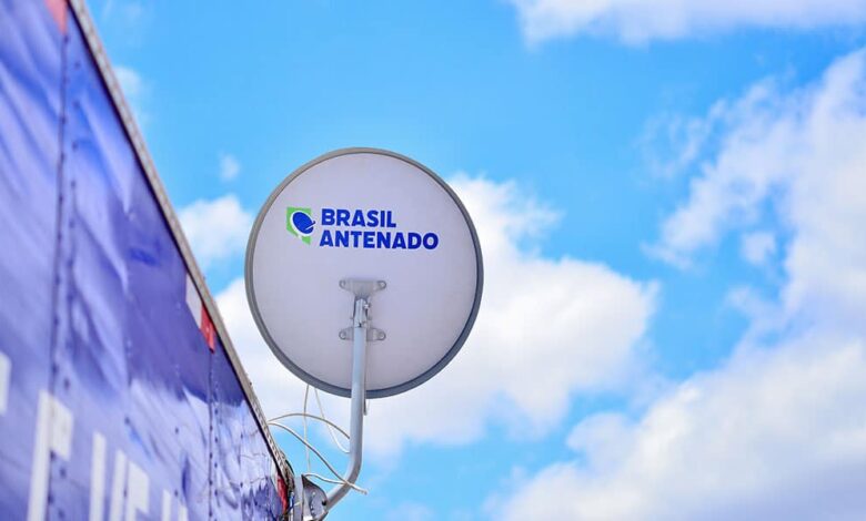 Antena parabólica digital branca com logotipo "Brasil Antenado" instalada em estrutura azul contra céu azul com nuvens.