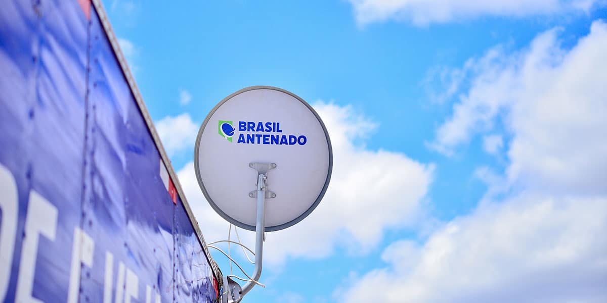 Antena parabólica digital branca com logotipo "Brasil Antenado" instalada em estrutura azul contra céu azul com nuvens.