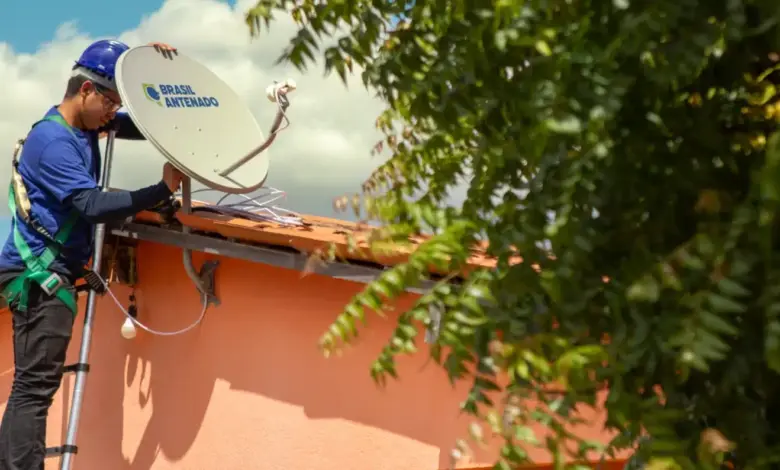 Técnico com capacete azul instala antena parabólica digital do programa Brasil Antenado no telhado de casa cor-de-rosa.