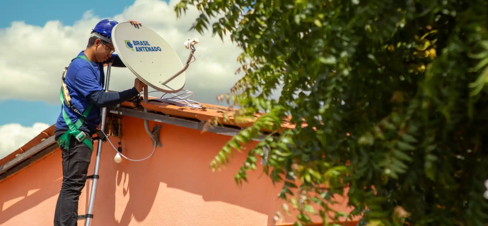 Técnico com capacete azul instala antena parabólica digital do programa Brasil Antenado no telhado de casa cor-de-rosa.