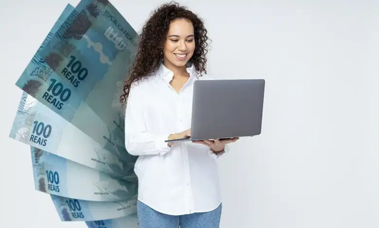 Mulher jovem sorrindo segura notebook cinza com notas de 100 reais ao fundo em fundo branco.