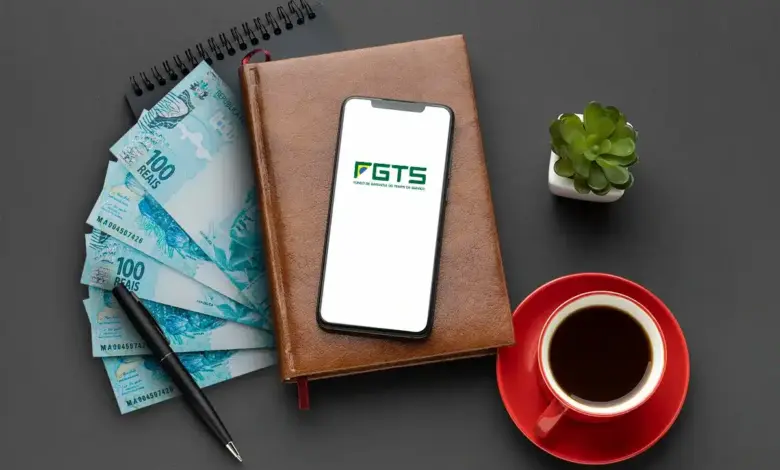 Celular exibindo logo do FGTS sobre caderno marrom, notas de R$ 100, caneta preta, xícara vermelha e suculenta.