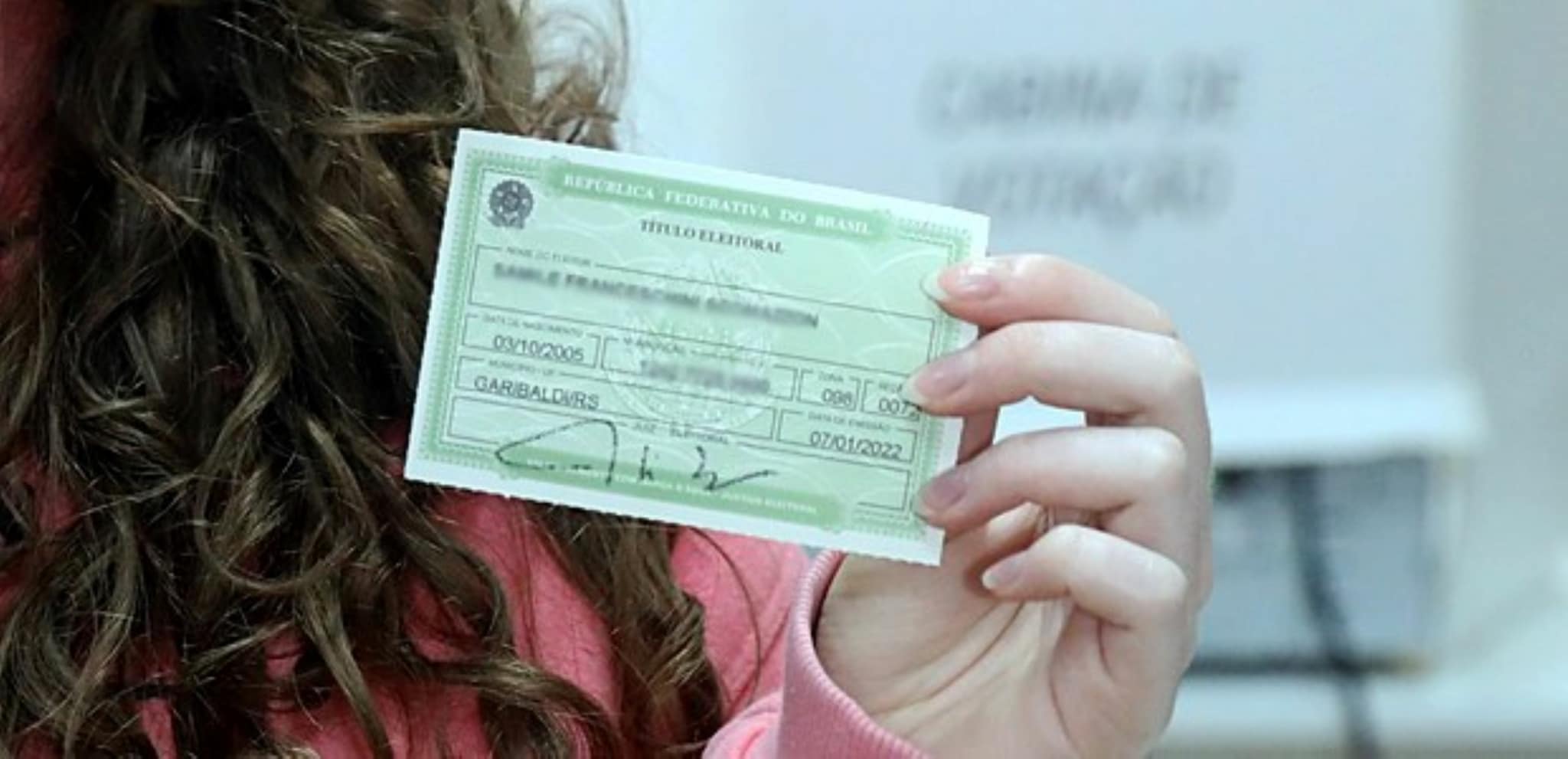 Mulher de cabelos castanhos cacheados segura título eleitoral verde da República Federativa do Brasil.