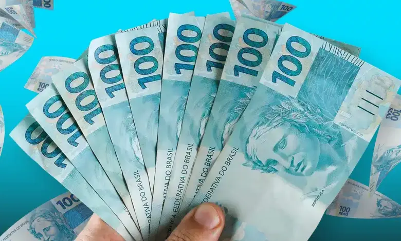 Mão segurando leque de notas de R$ 100 com fundo azul claro e cédulas espalhadas.