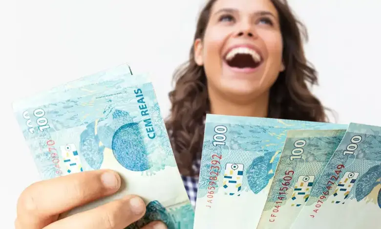 Mulher sorridente de cabelos castanhos ondulados segura quatro notas de R$ 100 em leque sobre fundo branco.