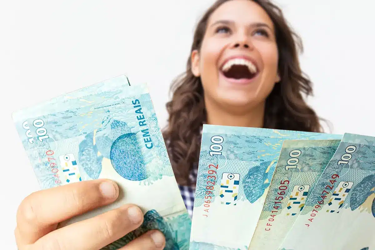 Mulher sorridente de cabelos castanhos ondulados segura quatro notas de R$ 100 em leque sobre fundo branco.